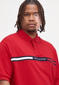 Rød polo T-shirt lavet af bomuld, med krave, tre knapper og en vandret stribe med marineblå og hvide accenter, der viser "TOMMY HILFIGER."