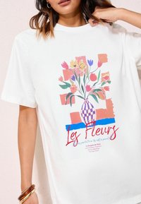 Weißes Baumwoll-T-Shirt mit einem bunten Blumenvase-Design in blau-orange Akzenten und dem Text "Les Fleurs" in fetten roten Buchstaben.