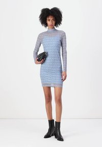 Langemouwige, lichaamssluitend, doorschijnende blauwe jurk met "HUGO" logo patroon; gestyled met een zwarte tas en enkellaarsjes.