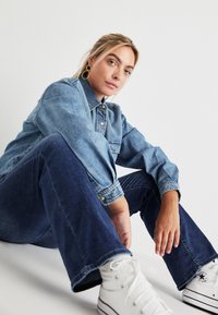 Chaqueta de mezclilla en azul claro, con un cierre de botones en la parte delantera y un corte relajado. Combinada con jeans azul oscuro y zapatillas blancas de caña alta.