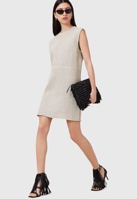 Knelange beige perforeret kjole med ærmeløst design. En sort frynse-clutch og remsandaler med hæl komplementerer outfittet.