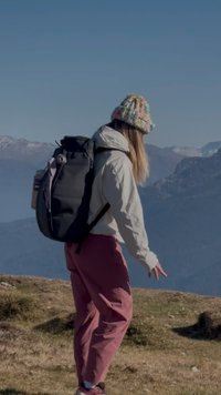 Pessoa com um gorro colorido, jaqueta branca e uma grande mochila preta a estar em terreno montanhoso coberto de relva, com picos nevados ao fundo sob um céu limpo.