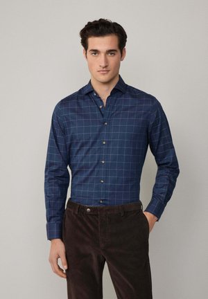Camisa de cuadros en tonos azul marino, hecha de una mezcla de algodón, con cierre de botones, cuello puntiagudo y mangas largas; combinada con pantalones marrones oscuros.