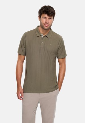 Hombre con camiseta polo de manga corta color verde oliva y pantalones beige, de pie con una mano en el bolsillo contra un fondo claro y sencillo.