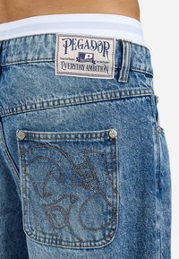 Blue denim shorts feature a woven label reading "PEGADOR" with decorative stitching on the back pocket. White waistband visible above.