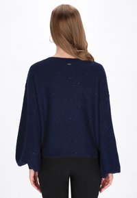 Marineblauwe gebreide trui met oversized mouwen, voorzien van een gestructureerd patroon en subtiele glittertjes. Ribbelband aan de taille.