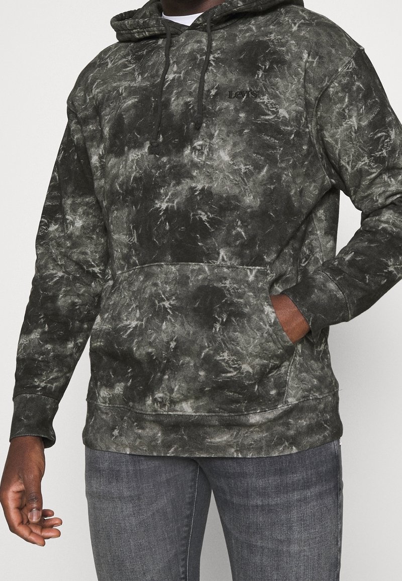 Homme portant un sweat à capuche Levi's noir et gris tie-dye avec poche avant et jean gris, main partiellement dans la poche, sur un fond uni.