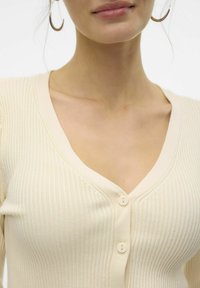Krämfärgad ribbad cardigan med V-hals, med tre knappar fram och subtil struktur. Smoother, lättviktig tyg.