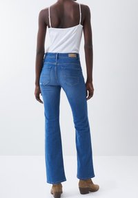Vaqueros de mezclilla acampanados de color azul, con cintura alta, dos bolsillos traseros y una etiqueta marrón en la cinturilla. La modelo lleva una camiseta sin mangas blanca.