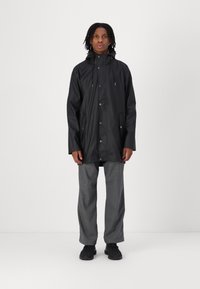 Black waterproof coat with a hood, featuring snap buttons and two side pockets, worn over gray pants and black shoes. Simple design.