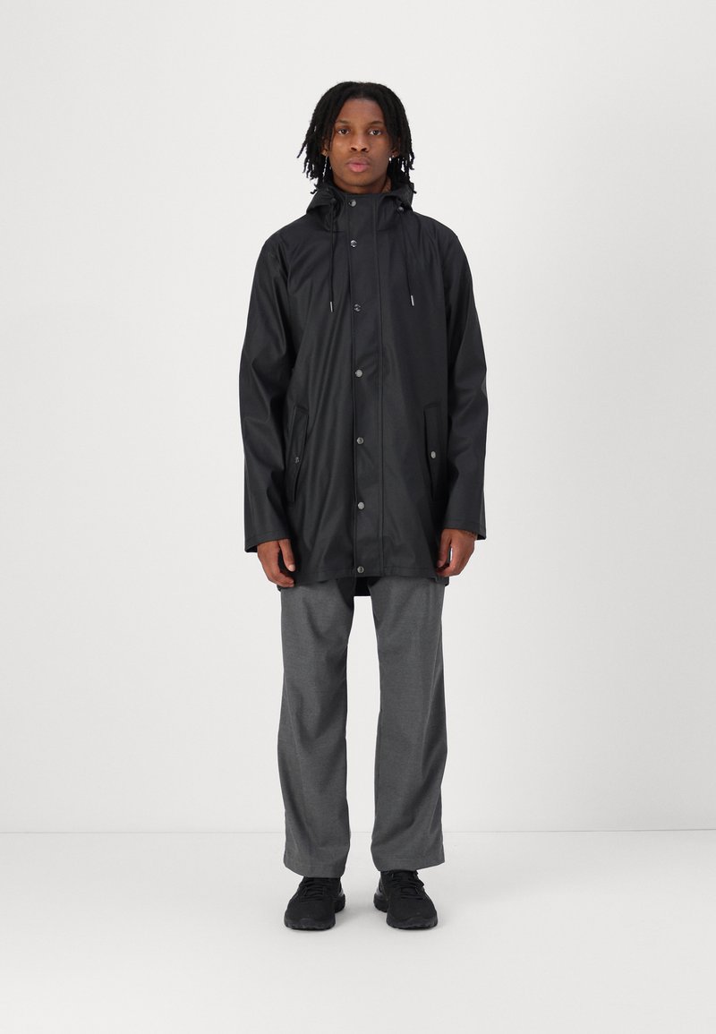 Black waterproof coat with a hood, featuring snap buttons and two side pockets, worn over gray pants and black shoes. Simple design.