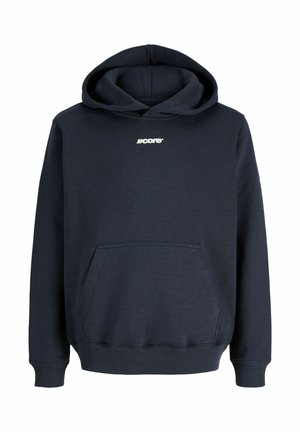Jongen met een zwarte pet, zwart jack, donkere hoodie, losse olijfgroene broek en zwarte sneakers staat met ontspannen handen en kijkt naar rechts.