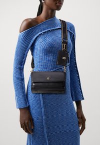 Bolso cruzado de cuero negro con un logo dorado, acoplado a un vestido azul acanalado con un patrón que presenta detalles de botones en las mangas y el costado.