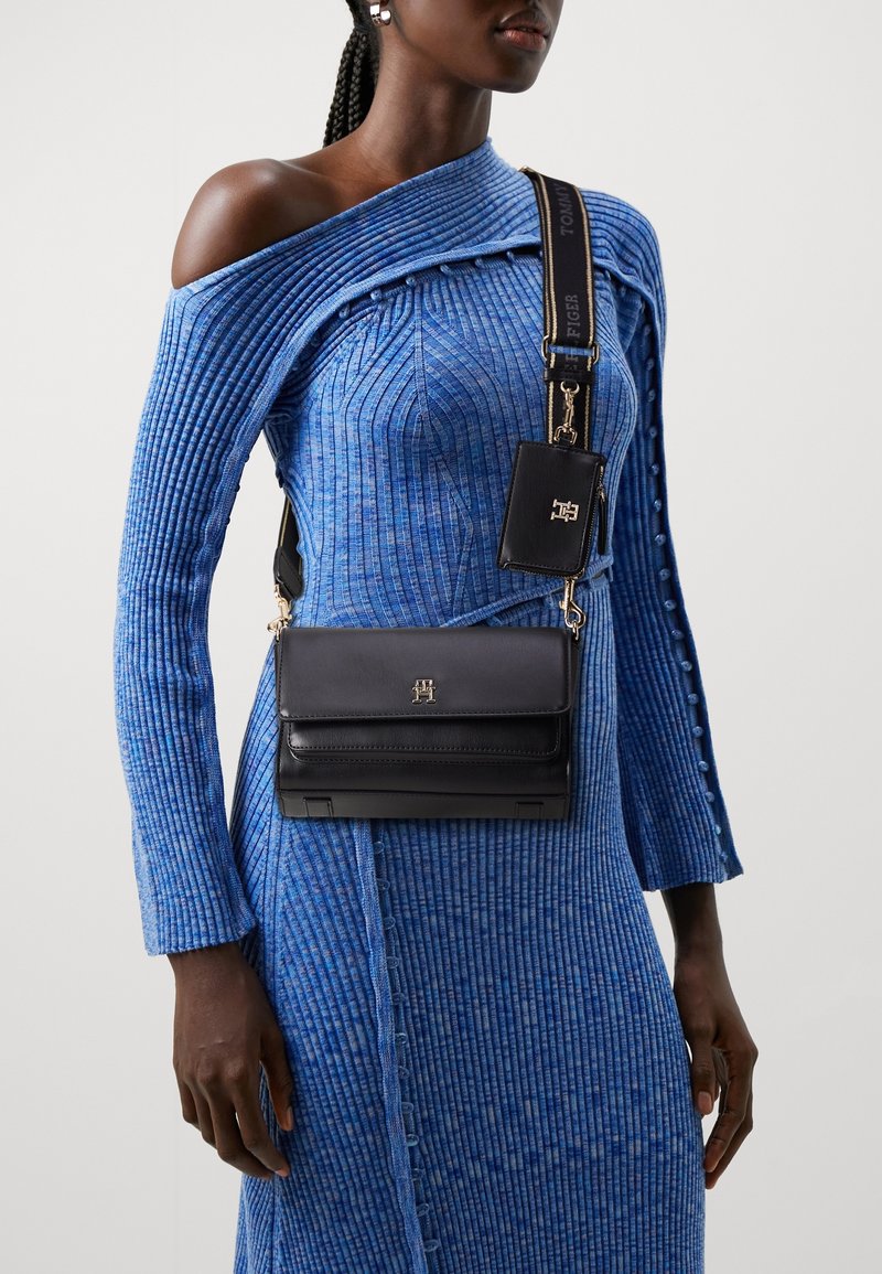 Bolso cruzado de cuero negro con un logo dorado, acoplado a un vestido azul acanalado con un patrón que presenta detalles de botones en las mangas y el costado.