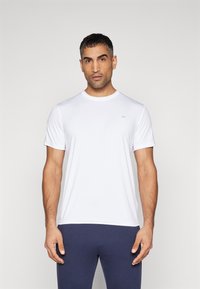 T-shirt branco de gola redonda, feito de tecido leve, com mangas curtas e um pequeno logótipo no peito. Usado com calças azul-marinho.