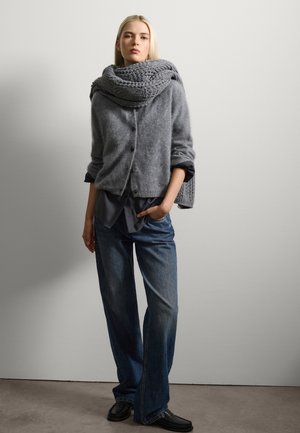 Gilet en tricot gris avec des boutons, superposé sur une chemise sombre. Un jean en denim à jambes larges et des chaussures noires à enfiler complètent le look.