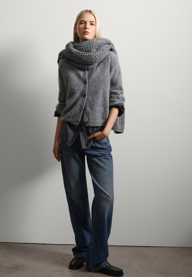 Gilet en tricot gris avec des boutons, superposé sur une chemise sombre. Un jean en denim à jambes larges et des chaussures noires à enfiler complètent le look.