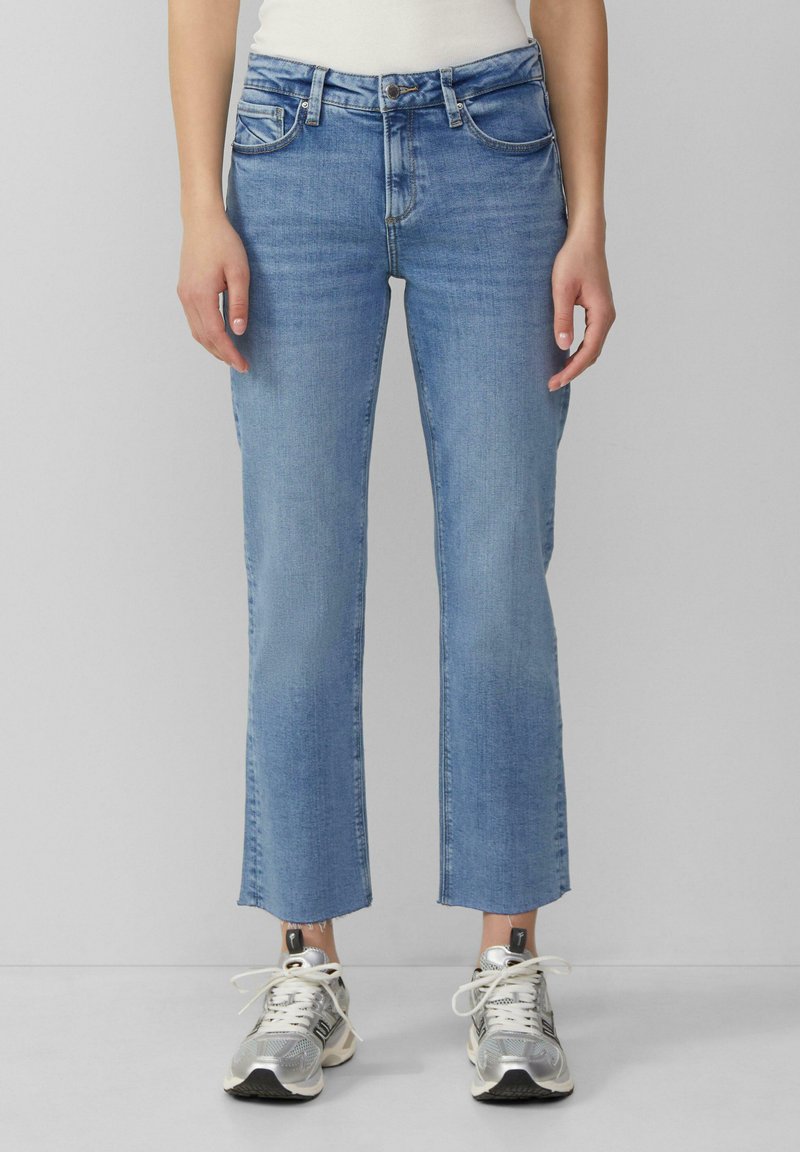 Jean en denim bleu à coupe droite, avec des ourlets raccourcis, cinq poches et une texture délavée. Porté avec des chaussures de sport argentées.