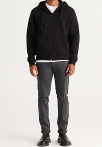 Zwarte hoodie met rits, voorzien van een trekkoord, kangoeroezakken en ribgebreide manchetten, gedragen over een wit T-shirt, gecombineerd met grijze broek en zwarte laarzen.