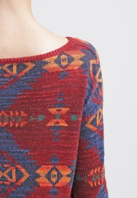 Strickpullover in Rot, Blau und Orange mit geometrischen Mustern. Weite Halslinie, weiche Textur und entspannte Passform erkennbar.