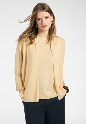 Mujer con cabello suelto castaño claro, vestida con un cárdigan beige abierto y pantalones oscuros, de pie con las manos en los bolsillos contra un fondo liso.