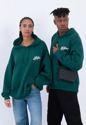 Sweat-shirts à capuche verts avec poches avant et cordons, arborant un logo blanc. Le mannequin masculin porte un sac en bandoulière noir. Jeans décontractés et pantalons.