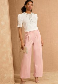 Weißes Oberteil mit kurzen Puffärmeln und Schleifendetail, kombiniert mit hellrosa, plissierten weiten Hosen. Hält eine kleine goldene Clutch.