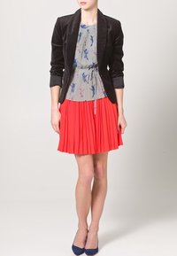 Černý sametový blejzr, květinová šedá blůza a plisovaná červená sukně. Oblečení má přiléhavý střih a různé textury s modrými podpatky.