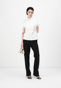 White short-sleeve blouse with ruffled front, paired with black tailored pants and black strappy heels. Model holding a small beige handbag.