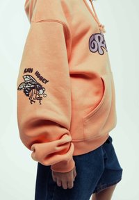 Sudadera melocotón con un bolsillo frontal, mangas oversized y diseños gráficos que presentan una abeja y "RAW HONEY" en la manga izquierda.