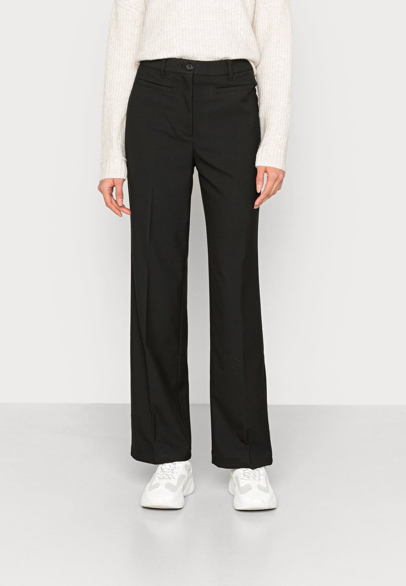 Femme portant un pantalon droit noir, un pull en maille crème à manches longues et des baskets blanches, debout devant un fond uni.