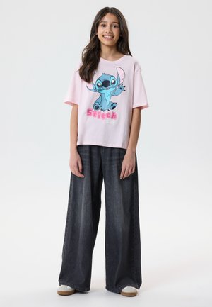 Młoda kobieta uśmiechająca się, ubrana w różowy T-shirt z niebieską grafiką Stitcha oraz szerokie czarne spodnie dżinsowe, stojąca na białym tle.