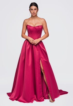 Femme aux cheveux foncés portant une robe bustier fuchsia longue jusqu'au sol avec une fente haute à la cuisse et des sandales argentées à talons hauts, debout, mains jointes.