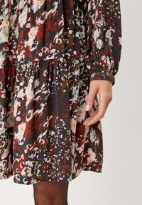 Blommönstrad klänning i svart, röd och teal, med en lager-på-lager kjol, långa ärmar och elastiska manschetter. Mjuk tyg med avslappnad passform.