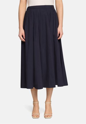 Femme portant une jupe midi plissée taille haute bleu marine associée à des escarpins beiges à bride à la cheville et bout ouvert sur fond blanc.