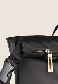 Mochila negra con una superficie texturizada, que cuenta con un bolsillo con cremallera y una correa resistente. Incluye una etiqueta con el nombre de la marca "FREDDY."