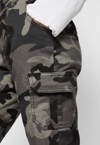 Pantalones cargo de camuflaje en tonos grises, con grandes bolsillos laterales, una textura suave y un corte relajado, con detalles de costura visibles.