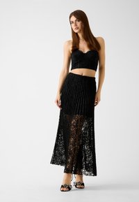 Top corto in pizzo nero con scollo a cuore abbinato a una lunga gonna nera trasparente plissettata. Scarpe rifinite con dettaglio a fiocco.