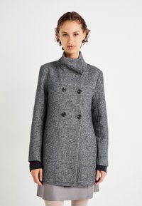 Manteau double boutonnage gris texturé avec col haut, boutons noirs et poignets noirs contrastants. Le tissu semble chaud et robuste.