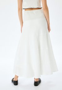 Jupe maxi blanche à volants présentant un tissu texturé et une taille élastique. Portée avec des sabots noirs, présentée sur un fond blanc.