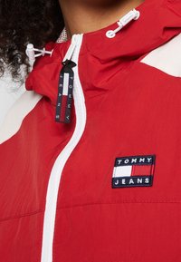 Czerwona kurtka wiatrówka z białymi akcentami i naszywką z logo. Projekt obejmuje zamek błyskawiczny i regulowany kaptur ze sznurkiem.