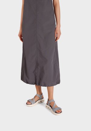 Persona che indossa un vestito grigio lungo fino alla caviglia e sandali imbottiti grigi con cinturini incrociati, in piedi su uno sfondo chiaro e uniforme.