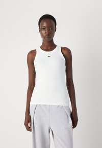 Hvit ribbet tank topp med rund hals, med en grønn brodert logo på brystet. Kombinert med grå joggebukser.