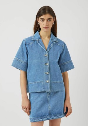 Lys blå denimskjorte med korte ærmer, spids krave og metalliske blomsterformede knapper. Har en firkantet pasform og pæne syninger.