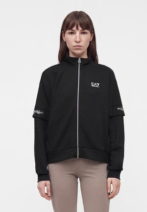 Kvinde iført en sort sportjakke med lynlås og EA7-logo på brystet samt broderi på ærmerne, kombineret med beige leggings, står foran en ensfarvet baggrund.