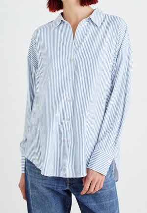 Kobieta ubrana w luźną, długorękawową koszulę na guziki w pionowe, jasnoniebiesko-białe paski oraz niebieskie dżinsy.