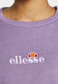 Detailní pohled na levandulovou mikinu s bílým logem "ellesse" a půlkruhovým oranžovým a červeným emblémem ve středu hrudníku.