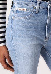 Šviesiai mėlyni džinsai su Calvin Klein etikete, artimiausias planas rodo penkių kišenių dizainą, lygų paviršių ir sidabrinius aksesuarus.
