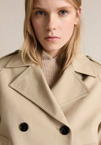 Beige dubbelknäppt trench coat med svarta knappar, breda slag och en texturerad inre krage, kombinerad med en ribbad neutral tröja.