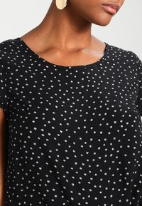 Czarny top z krótkimi rękawami, okrągłym dekoltem i rozproszonym białym geometrycznym wzorem na całej powierzchni. Gładka faktura i lekka tkanina.